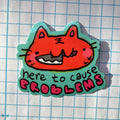 Sticker with a cartoon cat face and text 'here to cause problems' on a grid background