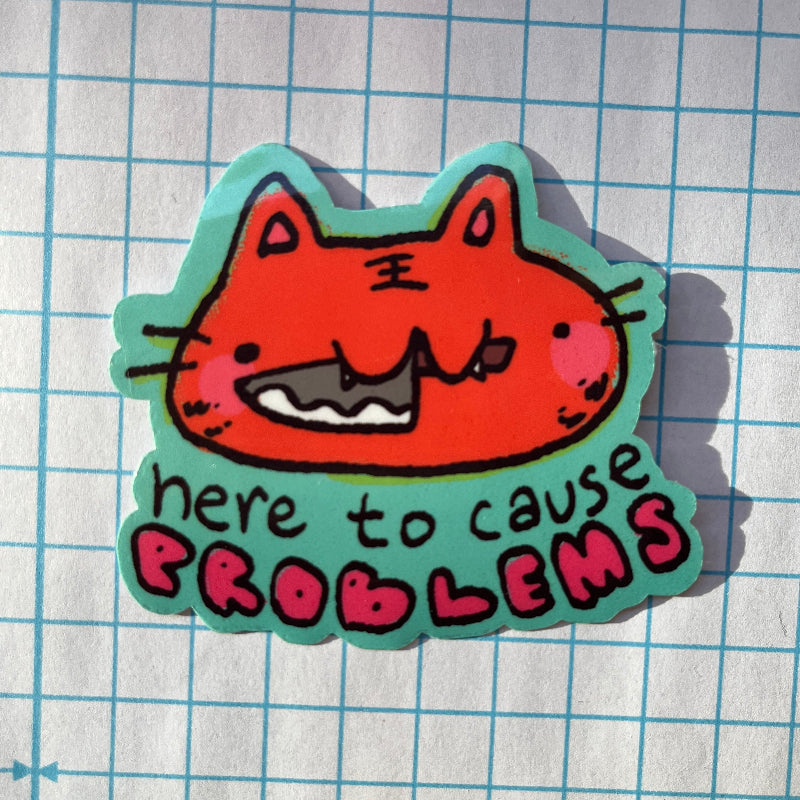 Sticker with a cartoon cat face and text 'here to cause problems' on a grid background